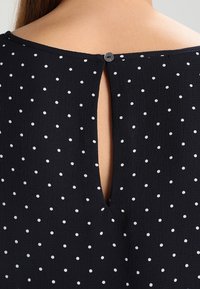 Černá blůza s puntíky, která má na zádech výřez ve tvaru klíčové dírky, zakončený kulatým knoflíkem. Měkká látka s texturovaným vzorem.
