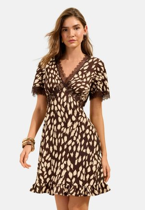 Junge Frau trägt ein braun-beiges Kleid mit Fleckenmuster, Spitzenbesatz, kurzen Ärmeln, Rüschenabschluss und goldenen Armbändern am Handgelenk.