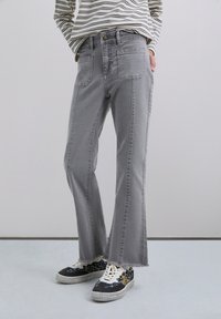 Jeans de mezclilla gris acampanados con un acabado desgastado, que presentan bolsillos frontales y traseros, y dobladillo sin rematar. Combinados con zapatillas de patrón.