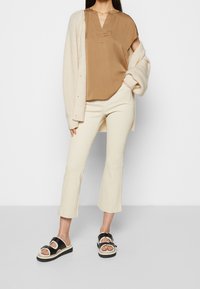 Beige Bluse mit V-Ausschnitt, cremefarbener Cardigan, beigefarbene ausgestellte Hose und schwarze Sandalen mit dicken Sohlen und silbernen Hardware-Akzenten.