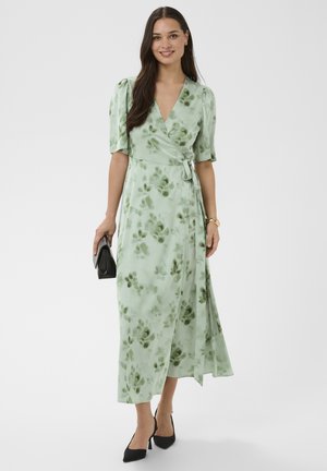 Donna che indossa un abito a portafoglio verde chiaro con motivi floreali e maniche fino al gomito, che tiene in mano una pochette nera e indossa tacchi neri e braccialetti dorati.