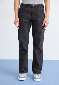 Pantaloni negri cu dungi fine, din material rezistent; dispun de buzunare frontale, închidere cu nasture și croi drept. Purtați cu adidași gri.