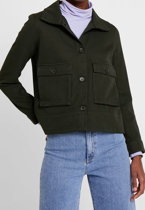 Schwarze, geknöpfte Jacke mit großen Fronttaschen, getragen über einen hellvioletten Rollkragenpullover, kombiniert mit hellblauen Jeans.