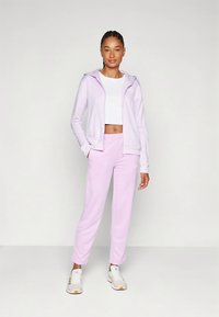 Svetlo vijolična zadrga z zadrgo in ujemajoče se trenirke iz mehke tkanine, v kombinaciji z belo kratko majico in belimi supergami.