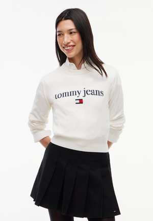 Kobieta o długich ciemnych włosach, ubrana w biały "tommy jeans" sweatshirt i czarną plisowaną spódnicę, uśmiecha się i patrzy na bok.