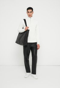Weißer Reißverschluss-Pullover, schwarze Lederhosen und eine große schwarze Tote-Tasche. Das Model trägt weiße Sneaker. Minimalistisches Design, glatte Texturen.