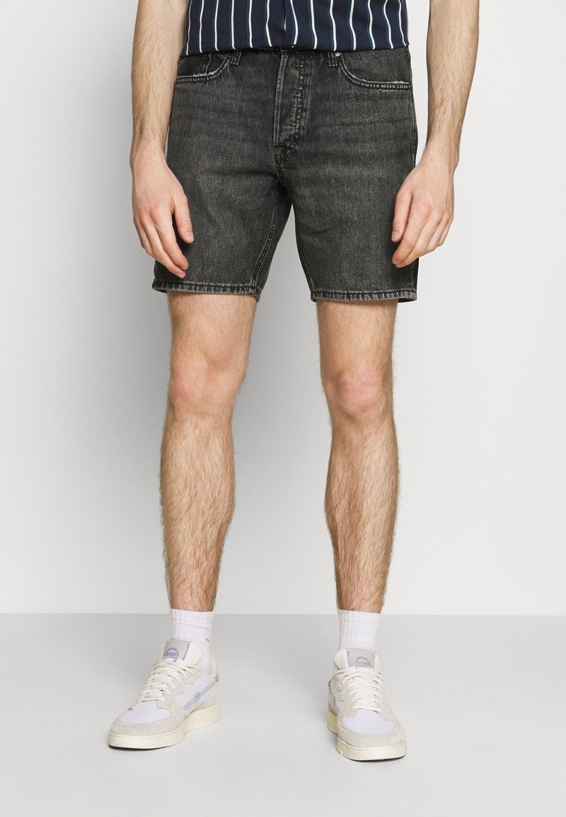 Shorts de mezclilla negros con diseño de cinco bolsillos, textura desgastada y dobladillo deshilachado. Combinados con zapatillas blancas y una camisa de rayas en azul marino.