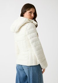 Giacca puffer bianca con cappuccio, dal texture trapuntato, con chiusura a zip e silhouette aderente. Abbinata a jeans di denim blu.