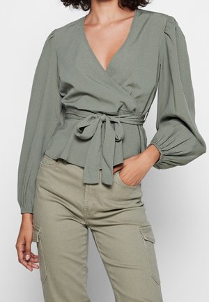 Blusa avvolgente verde, strutturata, con maniche lunghe a sbuffo e cintura con nodo. Abbinata a pantaloni cargo verde chiaro con tasche laterali.
