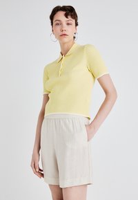 Gul ribbet polo skjorte med hvit kant, paret med lyse beige elastiske shorts. Modellen står foran en nøytral bakgrunn.