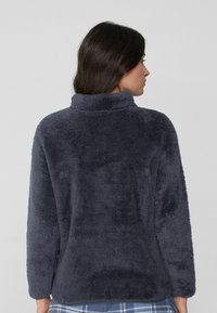 Suéter de polar gris oscuro con un cuello alto, con una textura suave y mangas largas, visto desde la parte posterior, combinado con pantalones estampados.