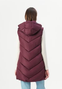 Gilet matelassé bordeaux avec une capuche, présentant un design matelassé en chevrons. Le dos a des coutures visibles et une coupe ample, associé à un léger pull.