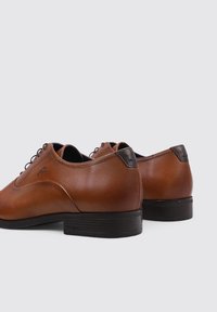 Chaussures en cuir marron avec une texture lisse, bout rond, accents noirs au talon et lacets ronds. Talon légèrement surélevé.