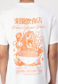 Hvit t-skjorte med oransje grafisk design av en løve og en ramenbolle, med tekst på engelsk og japansk, som viser sterke linjer og detaljerte mønstre.