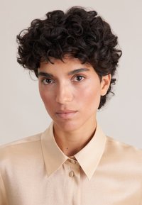 Camicia beige chiaro con una texture satinata liscia, caratterizzata da un colletto classico e chiusura con bottoni. Capelli scuri e ricci parzialmente visibili.