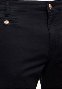 Gros plan sur un pantalon noir avec des boutons couleur cuivre, une poche avant visible, une boucle de ceinture et des coutures détaillées sur le tissu.