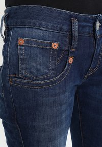 Jeans in denim blu scuro presentano un classico design a cinque tasche, cuciture a contrasto, hardware in rame e una vestibilità aderente in vita.