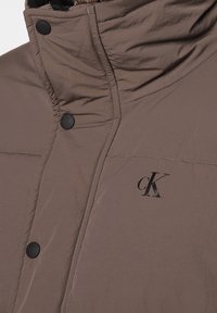 Brun vadderad jacka med hög krage, svarta tryckknappar och en subtil cK-logotyp på tyget. Slät och lätt textur.