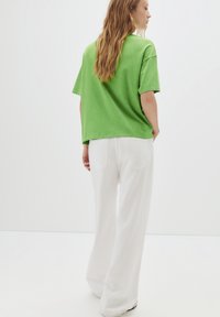 T-shirt oversize verde realizzata in tessuto morbido, abbinata a pantaloni bianchi a gamba larga. Texture liscia, vestibilità casual e senza motivi o decorazioni visibili.
