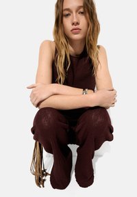 Burgundowy, teksturowany sweter połączony z luźnymi spodniami z wyraźnymi troczkami i frędzlami. Model siedzi w zrelaksowanej pozycji.