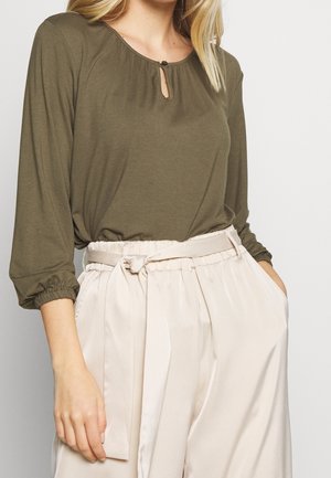 Femme portant un haut à manches longues vert olive avec un col montant à bouton en forme de goutte, associé à un pantalon taille haute crème style paperbag avec une ceinture en tissu.