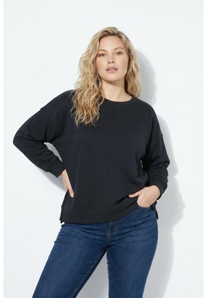 Kvinde med langt, bølget blondt hår iført en sort sweatshirt og blå jeans, stående med den ene hånd i lommen mod en hvid baggrund.