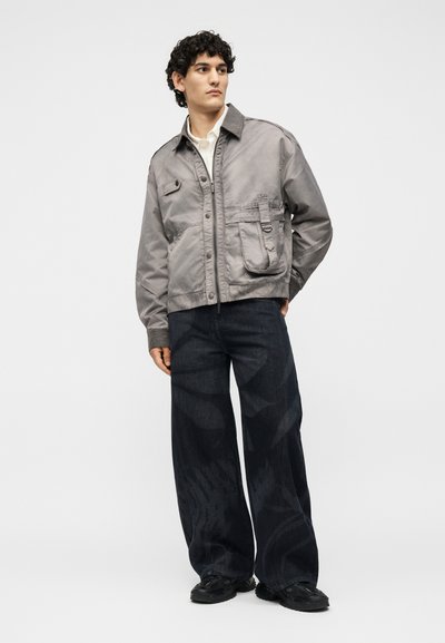 Νέος άνδρας με σγουρά μαλλιά που φοράει γκρι σακάκι με κουμπιά, φαρδιά σκούρα παντελόνια με σχέδια, μαύρα παπούτσια, στέκεται μπροστά σε απλό λευκό φόντο.