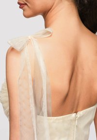 Épaule et haut du dos d'une femme portant une robe crème clair avec un nœud en tulle transparent noué sur une épaule.