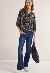 Jeune femme souriante en blouse fleurie, jeans évasés bleus, baskets blanches, tenant une veste matelassée noire sur un fond uni.