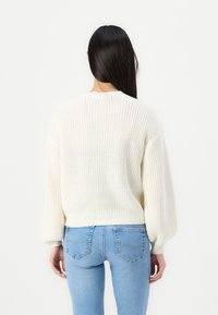 Un pull crème en tricot épais avec des manches bouffantes, un ourlet côtelé et des détails de couture au dos, associé à un jean ajusté bleu clair.