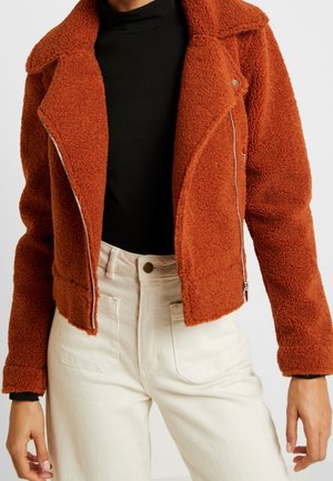 Rostfarbene, strukturierte Zip-Up-Jacke über schwarzem Top, kombiniert mit hoch taillierten, cremeweißen Hosen mit Fronttaschen, gezeigt an einer stehenden Person.