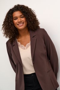 Blazer marron en tissu lisse, avec un bouton unique, deux poches avant et une coupe décontractée sur un top crème clair avec des détails en dentelle.