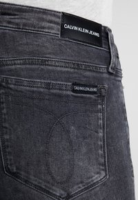 Jeans de mezclilla gris con una etiqueta de marca de cuero negro en la cintura y una pequeña etiqueta en el bolsillo trasero, con detalle de costura curva.