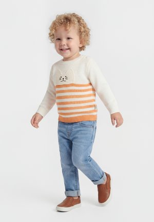 Peuter met krullend haar draagt een oranje gestreepte trui met kattenprint, lichtblauwe spijkerbroek en bruine instapschoenen, lopend en glimlachend.