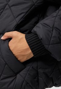 Must mustriline tõmbluku ja ribide äärega musta värvi jakk, millel on käsi küljetaskus, rõhutades tekstuurilist materjali ja õmbluste detaile.