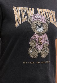 Μπλούζα με φθαρμένο τύπωμα αρκουδάκι με ροζ καπέλο και μπλούζα, με τα γράμματα "K", "NEW YORK" πάνω και τη φράση "ONE TEAM, ONE HEARTBEAT" κάτω.