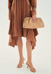 Kobieta w brązowej plisowanej asymetrycznej sukience trzymająca beżową marszczoną kopertówkę, ubrana w beżowe sandały na wysokim obcasie z paseczkami na jasnym tle.