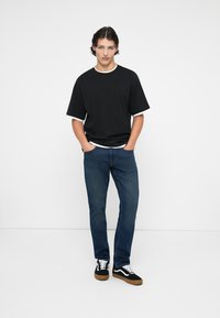 Camisa negra de manga corta con ribete blanco, combinada con jeans de mezclilla azules y zapatillas negras con detalles blancos. Pose de pie contra un fondo liso.