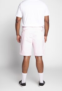 Světle růžové bavlněné šortky se zadními kapsami a zlatými knoflíky, kombinované s jednobarevným bílým tričkem a černými teniskami.