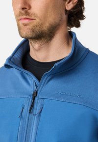 Blaue Zip-Jacke mit hohem Kragen, strukturiertem Stoff und zwei Reißverschlusstaschen. Mit gesticktem Logo auf der Brust.