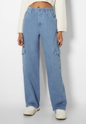 Personne portant un pantalon cargo taille haute bleu clair avec des poches latérales et des baskets blanches, associé à un haut blanc à manches longues raccourci.