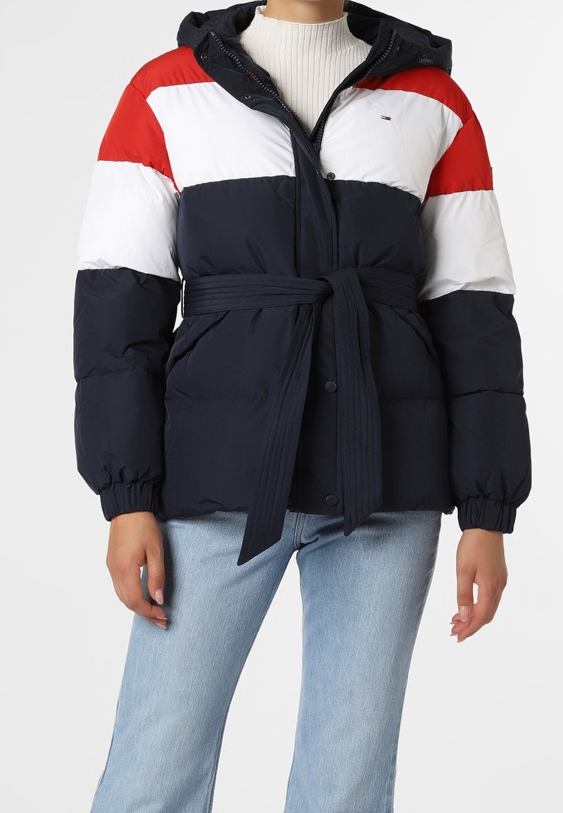 Veste en duvet bleu marine avec des rayures horizontales rouges et blanches, une ceinture à la taille et une capuche. Comprend des poignets élastiques et des boutons.