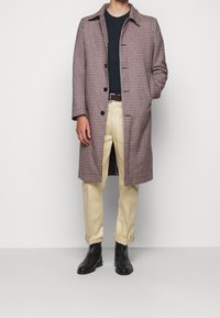 Hnědo-bílý kostkovaný kabát s plochým límečkem, na knoflíky a bočními rozparky, doplněný černými botami a krémovými kalhotami.