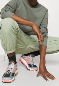 Scarpe sportive grigie con accenti arancioni, tomaia in mesh strutturato, suola ammortizzata, abbinate a pantaloni joggers verde chiaro e a una felpa grigia coordinata.