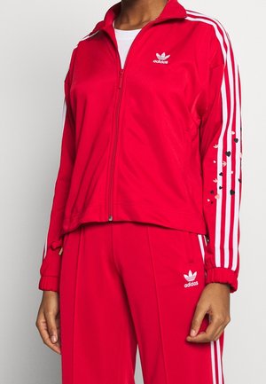 Personne portant un survêtement rouge Adidas avec des bandes blanches et de petits motifs en forme de cœur sur la manche, debout devant un fond uni.
