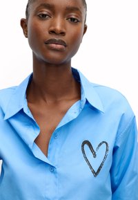 Jasnoniebieska koszula zapinana na guziki z czarnym wzorem serca po lewej stronie klatki piersiowej. Gładki bawełniany materiał z klasycznym projektem kołnierzyka.