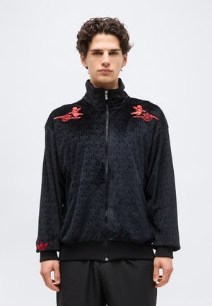 Joven con cabello rizado que lleva una chaqueta negra con cremallera con bordados de querubín rojo y cañón en los hombros y un patrón repetido de "AFC".