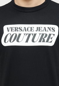 Μαύρο t-shirt με λευκό ορθογώνιο λογότυπο που γράφει «VERSACE JEANS COUTURE» με έντονα μαύρα κεφαλαία γράμματα.