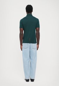 Polo shirt scurt, de culoare verde închis, cu guler, asortat cu blugi decolorati, lejeri, de culoare albastră deschisă și pantofi negri, văzut din spate.