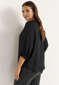 Svart, lätt blus med avslappnad passform, som har trekvartslånga puffärmar och en krökt fåll. Materialet har en slät yta.
